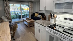 Cocina compacta con armarios blancos, microondas y estufa. Hay una zona de comedor con mesa y sillas, además de un sofá con cojines.