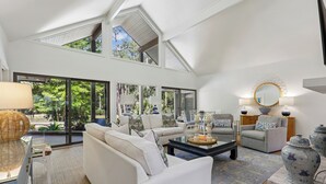 A spacious living room with a high ceiling, large windows, and a view of greenery.