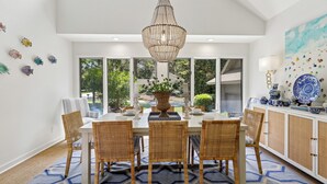 A dining area with a chandelier, wicker chairs, a table set for four, and a view of greenery outside.
