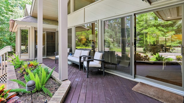 A covered outdoor deck with a seating area, large windows, and a view of the surrounding greenery.