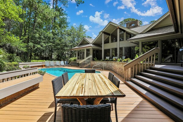 Una terraza de madera con piscina, mobiliario exterior y una escalera que lleva a un edificio con amplios ventanales.
