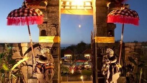 Une porte d’entrée traditionnelle, ornée de deux statues, surmontée d’un toit rouge et jaune, et offrant une vue sur une maison en arrière-plan.