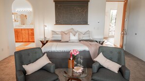 Une chambre avec un grand lit, deux fauteuils, une table basse en verre, une carte accrochée au mur, et une vue sur la cuisine et le balcon, visibles à travers une arche.