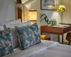 A neatly made bed with white linens and two decorative pillows with a leaf pattern. A wooden desk with a lamp and a plant is visible in the background. - Penh House Hotel (Phnom Penh)