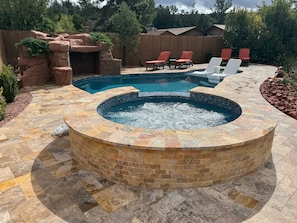 A circular hot tub with a brick surround, a swimming pool with a waterfall feature, and red lounge chairs.