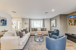 En moderne stue med sofa, lænestole og sofabord. Der er pejs, et maleri og et vindue med gardiner.