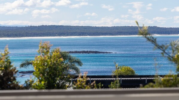 A coastal view with a clear sky, a sandy beach, and a forested hill in the distance.