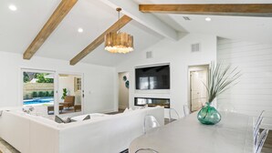 A modern living room with a large white sectional sofa, a dining table with chairs, a TV mounted on the wall, and a ceiling with exposed wooden beams.