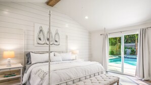 A bedroom with a four-poster bed, a bench, a nightstand with a lamp, and a view of a swimming pool through a glass door.