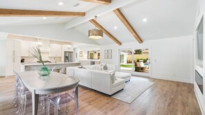 A spacious living area with a white sofa, clear plastic chairs, a dining table, and a kitchen in the background.