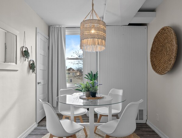 A modern dining area with a glass table, white chairs, a hanging light fixture, and a view of buildings outside. - Broad Street Suites 2 & 3 | Parking for 5 Vehicles |Downtown|Walkable |1 Block to Horticultural Hall (Lake Geneva)