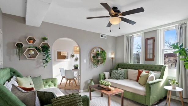 Un salon avec un canapé vert, une table basse et un ventilateur de plafond. Les murs sont ornés d’étagères hexagonales où sont disposées des plantes, et il y a une fenêtre avec des rideaux.
