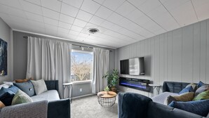 Un salon moderne comprenant un ensemble de canapés, une table basse, un téléviseur à écran plat fixé au mur et une fenêtre avec des rideaux.