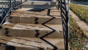 Une maison avec une porte d’entrée blanche, un petit porche et un escalier en béton doté de rampes en métal.