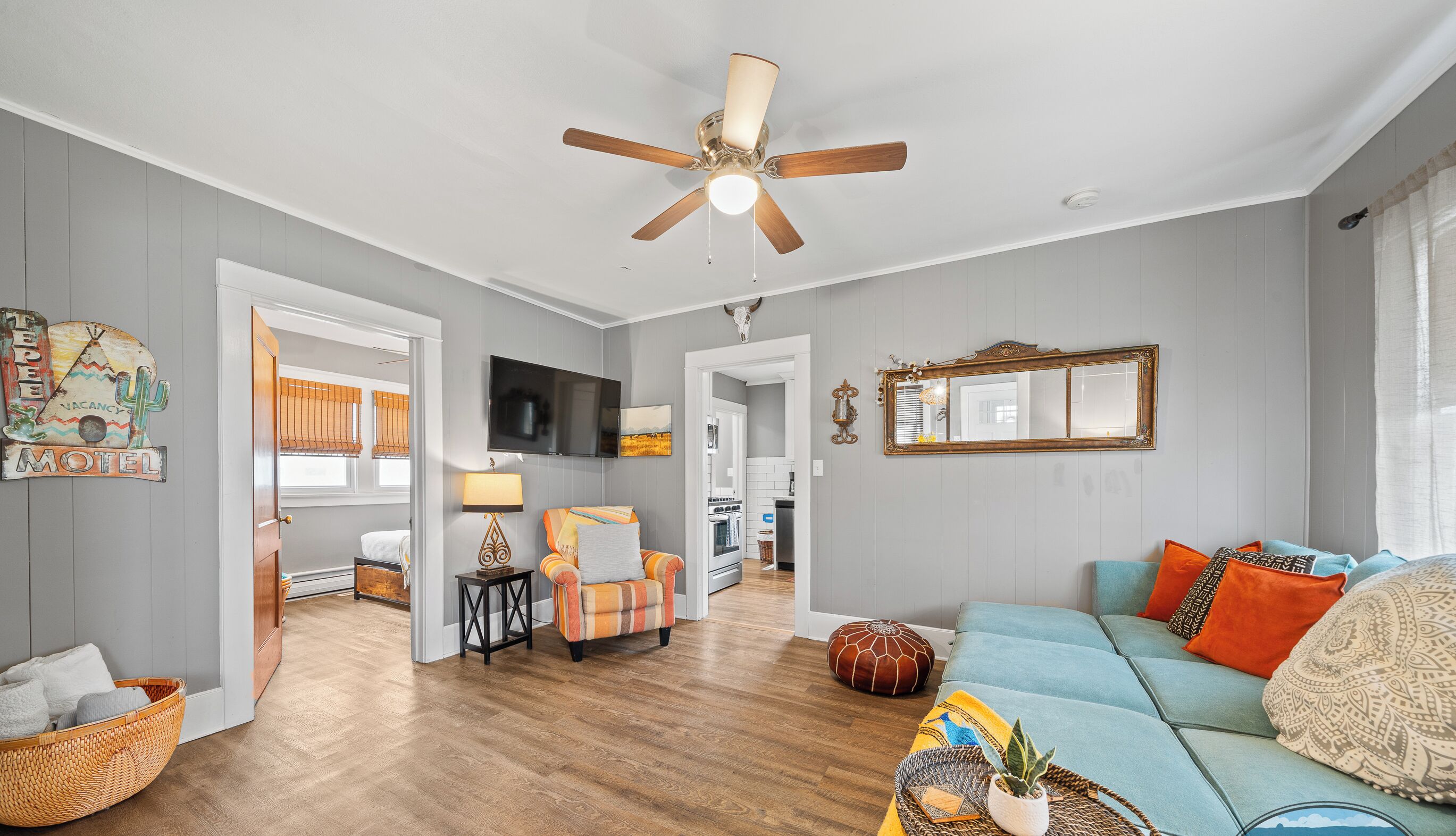 A spacious living room with hardwood flooring, a ceiling fan, a television, and a cozy seating area with a blue sofa and orange pillows.