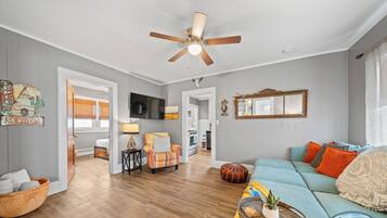 A spacious living room with hardwood flooring, a ceiling fan, a television, and a cozy seating area with a blue sofa and orange pillows.