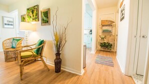 Una habitación con piso de madera, una silla de mimbre con cojín, un florero con ramas y una pared con cuadros enmarcados.
