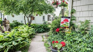 A garden pathway leading to a house with a 'Welcome' sign, surrounded by lush greenery and potted plants.