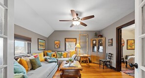 A cozy living room with a ceiling fan, a sofa with colorful cushions, a wooden coffee table, and a cabinet.
