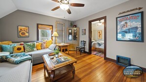 A cozy living room with a ceiling fan, a sofa with colorful cushions, a wooden coffee table, and a framed poster of the Titanic.