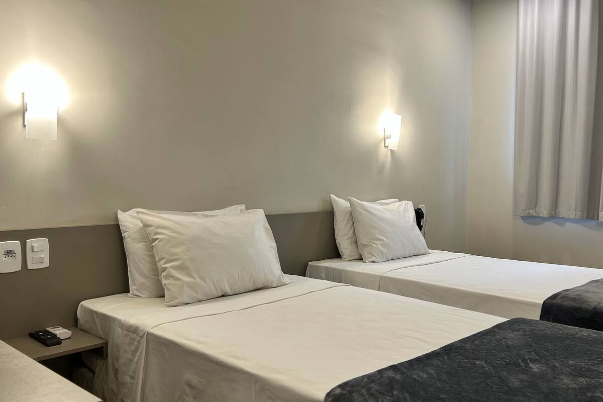 A hotel room with two beds, each with white bedding and pillows, a bedside table with a remote control, and a window with curtains.