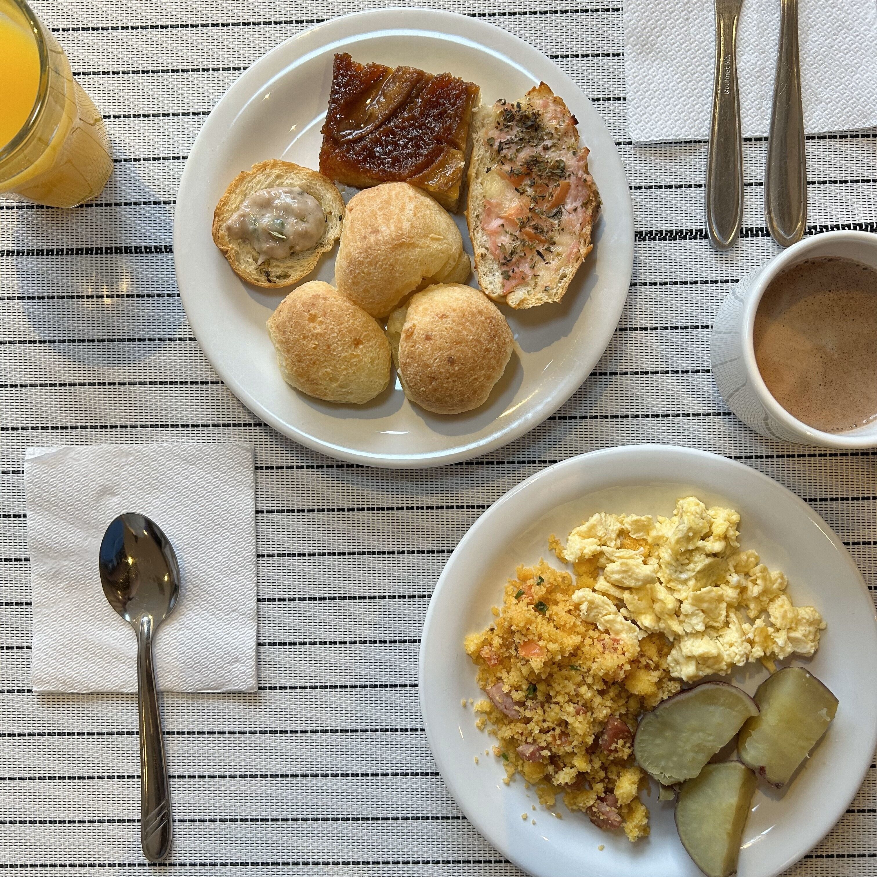Prato de café da manhã com ovos mexidos, batatas e pães em uma mesa com toalha listrada.