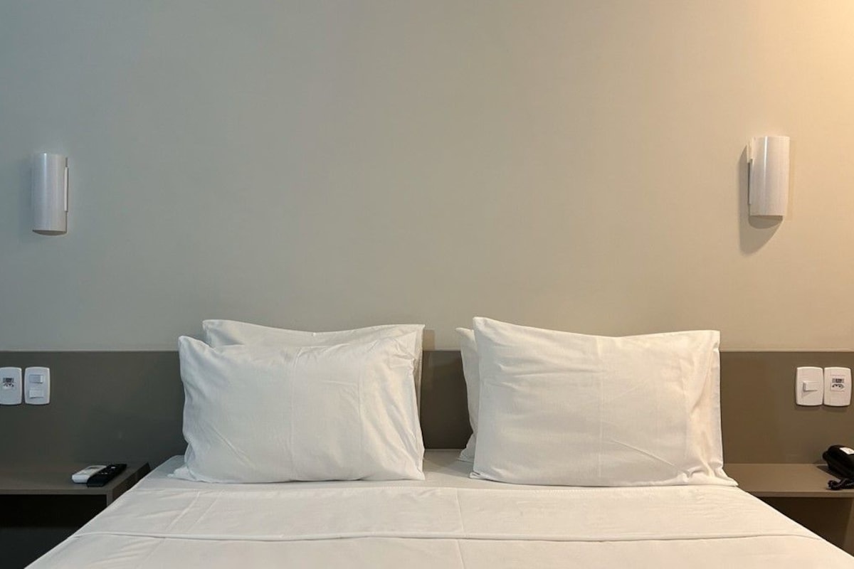A hotel room with a bed, two pillows, a bedside table with a phone, and a wall-mounted light fixture.