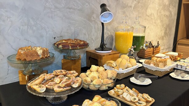 Una mesa de buffet con diversos alimentos, como pan, pasteles y un pastel, junto con un dispensador de jugo y una lámpara.