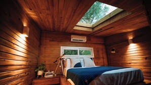 A cozy wooden cabin bedroom with a skylight, a bed, a bedside table with a lamp, and a small plant. - Vila Nambu (Gramado)