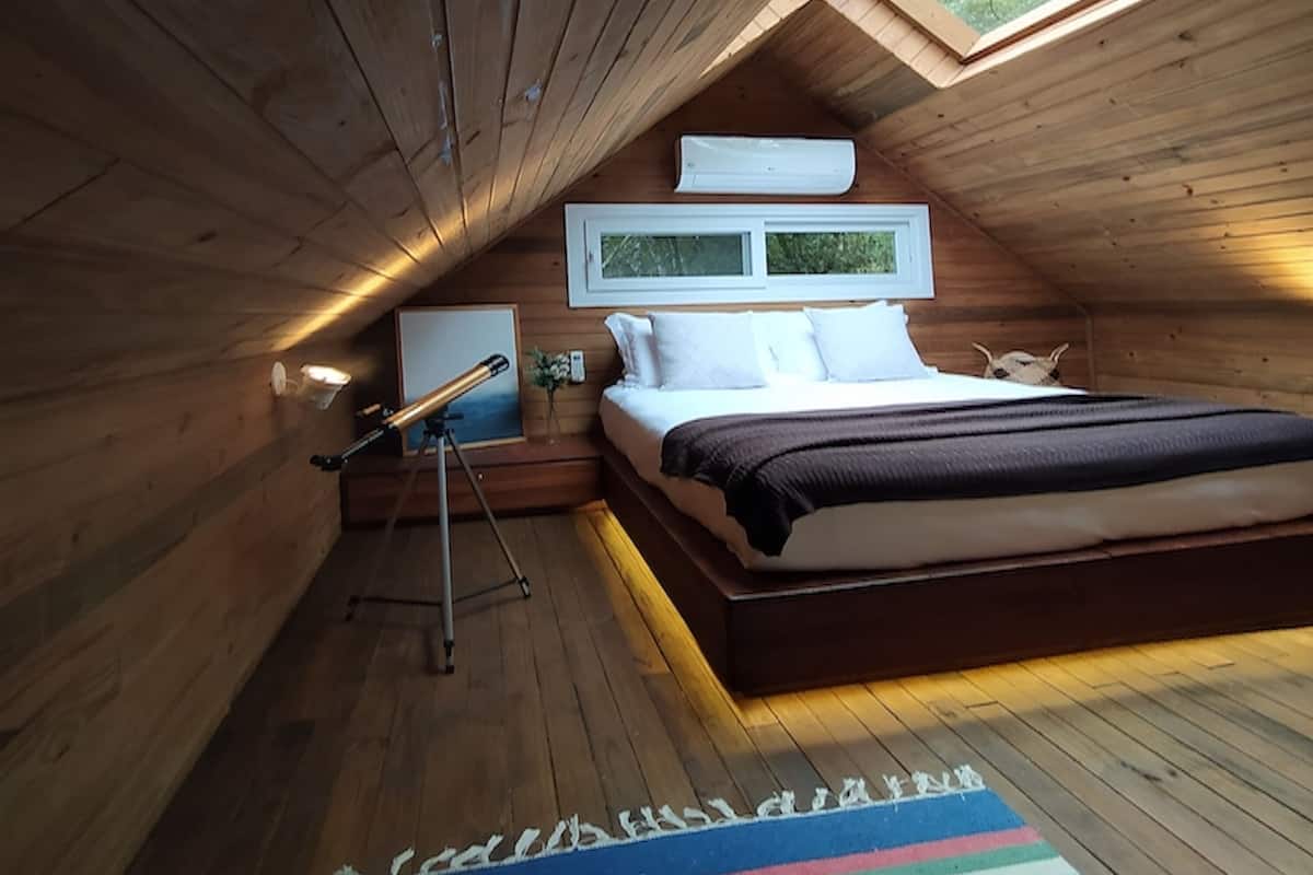 A wooden interior with a sloped ceiling, a bed, a telescope, and a window.