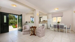 A spacious living area with a dining table and two armchairs, a kitchen with white cabinets, and a door leading to a garden. - Jasmine Luxury Villa With Private Pool (Corfu)
