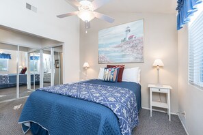 Une chambre à coucher avec un grand lit, un ventilateur de plafond, un tableau représentant un phare et une fenêtre avec des stores.