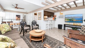 A spacious living area with a dining table, two wicker chairs, a round wooden coffee table, and a television displaying a beach scene. - Retro Split-Level Beach House | Steps to Sand | Darling Details Throughout! | Walk to Everything (Newport Beach)