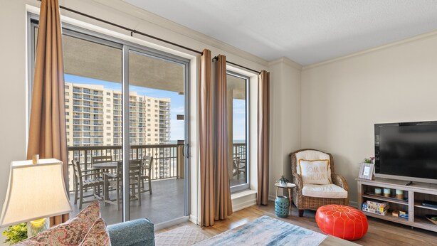 A modern living room with a sofa, a red ottoman, a TV, and a balcony with a view of a high-rise building. - $150 activities - E-gift card when you book - Balcony Access from Every Bedroom | Oceanview | MG1108 (Myrtle Beach)
