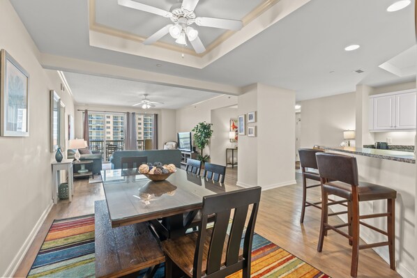 A spacious dining area with a wooden table and chairs, a ceiling fan, and a view of the living room and kitchen in the background.