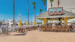 A beachfront restaurant with outdoor seating, palm trees, and boats in the background.