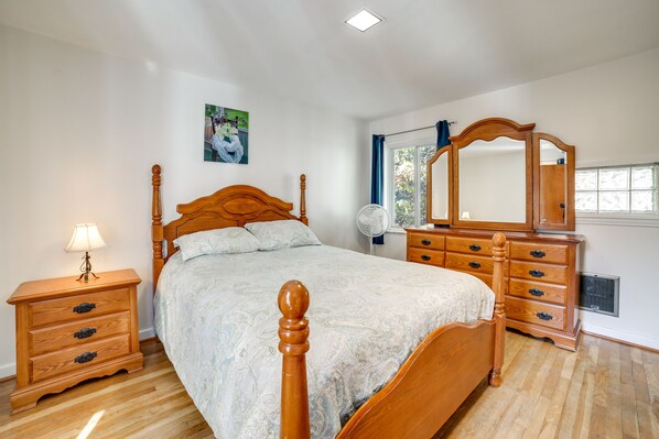 Quarto com cama de madeira, mesa de cabeceira com abajur, cômoda com gavetas, espelho e janela com cortinas.