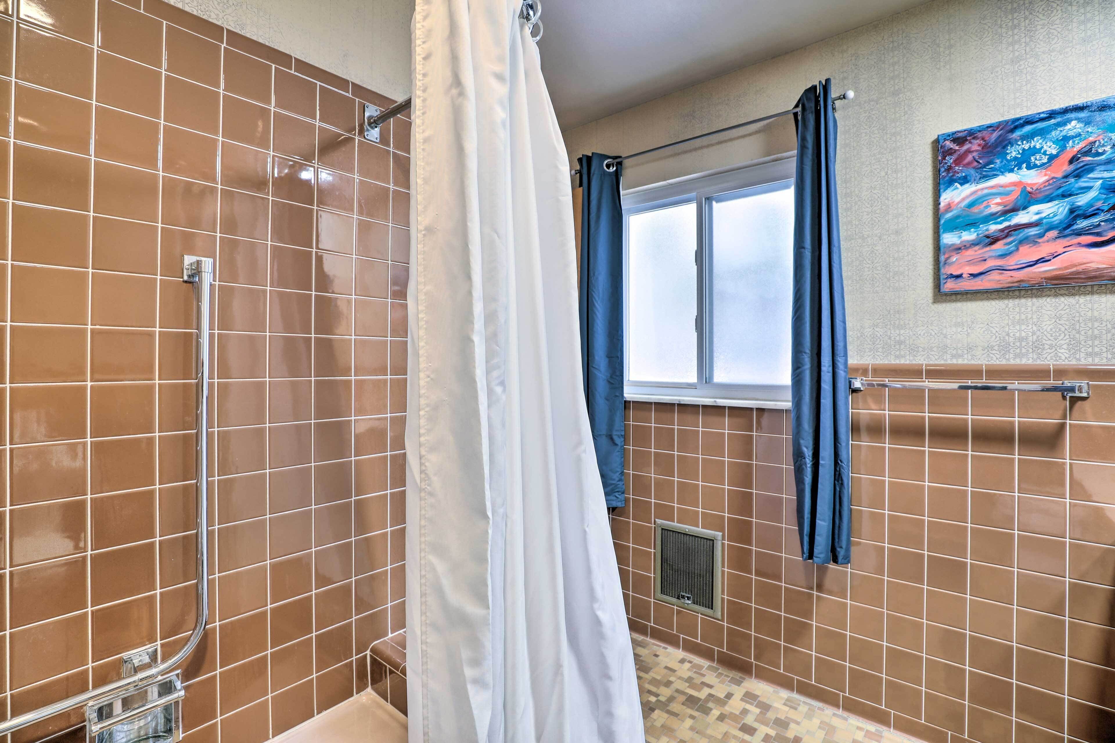 A bathroom with brown tiled walls, a shower with a curtain, a window with blue curtains, and a painting on the wall.