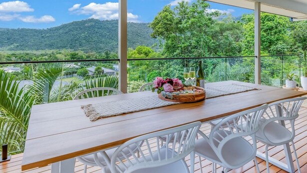 Un balcon avec une table en bois, des chaises blanches et une vue sur un paysage montagneux.