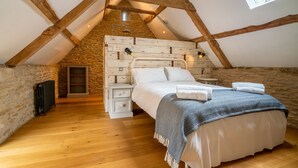 A bedroom with a sloped ceiling, exposed wooden beams, a stone wall, a wooden floor, a bed with white linens, a bedside table, and a wall-mounted lamp. - Middle Barn - sleeps 6 guests  in 3 bedrooms (Chippenham)