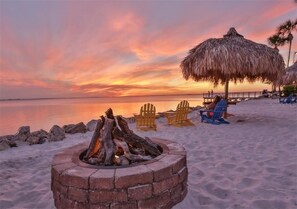 Eine Strandansicht mit einem Lagerfeuer, Liegestühlen und einem Reetdachschirm.