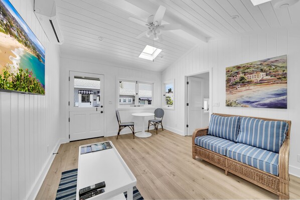 A room with a white ceiling, a ceiling fan, a dining table with chairs, a wicker sofa, and a painting of a beach scene.
