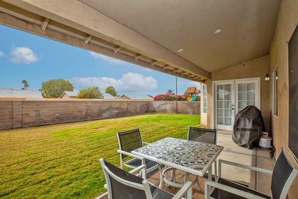 A patio with a table and chairs, a covered area, and a view of a backyard with a lawn and a brick wall.