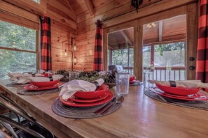 Une table à manger en bois, dressée avec de l’argenterie rouge, des verres et des couverts, et offrant une vue sur l’extérieur grâce à de grandes fenêtres.