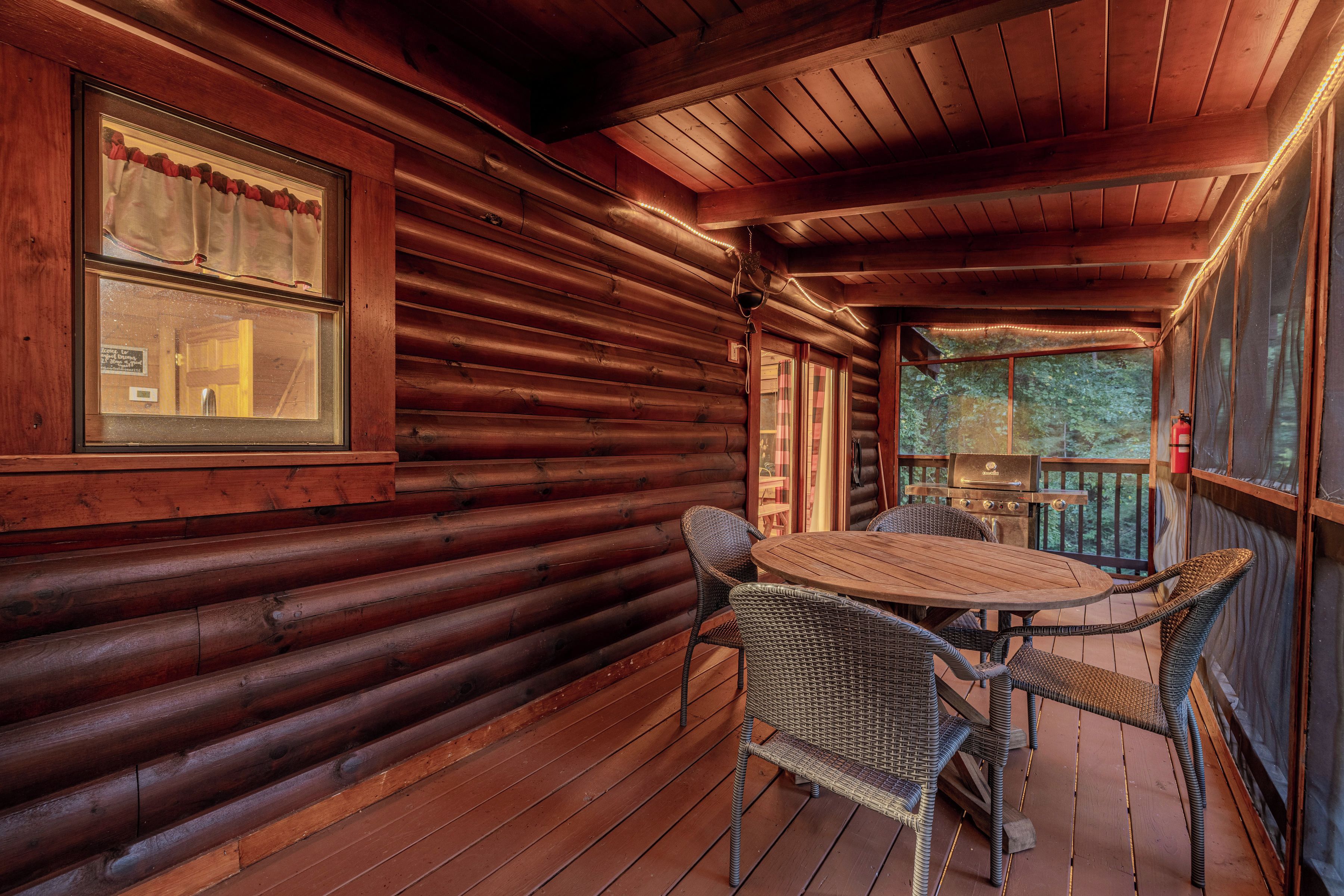 A wooden cabin with a covered porch, wicker chairs, a round table, and a grill.