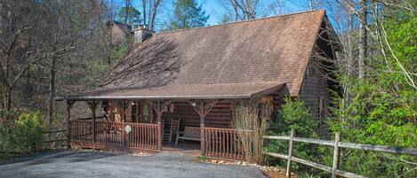 A rustic cabin with a porch, surrounded by trees and a wooden fence.