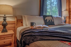 Una cama bien hecha con un libro, una taza y un almohadón con diseño de oso. Hay dos lámparas de noche y una mesita de noche de madera con un cajón.