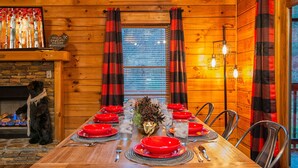 Ein Esszimmer mit Holzboden, einem langen Tisch, der für eine Mahlzeit gedeckt ist, gerahmt von roten und schwarzen Karosvorhängen, und einer Bärenfigur auf der linken Seite.