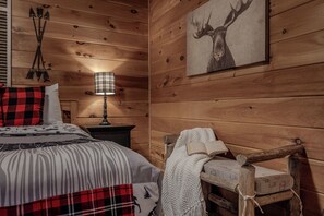 Ein Zimmer in einer Holzhütte mit einem Bett, einem Nachttisch, einer Lampe, einer Decke und einem Sessel.