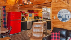 Ein Interieur einer Hütte mit einem roten Kühlschrank, Holzwänden, einem Esstisch, einem Ledersessel und einer Couch mit einem Karo-Kissen.